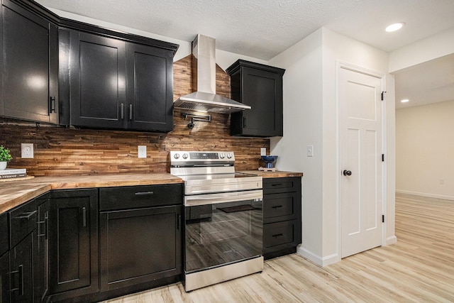 kitchen featuring dark cabinetry, stainless steel electric stove, wall chimney range hood, backsplash, and butcher block counters