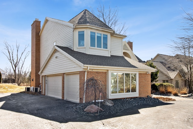 view of side of property featuring driveway, roof with shingles, an attached garage, brick siding, and a chimney