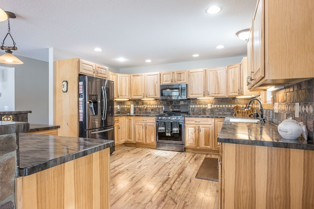 kitchen with light brown cabinetry, a sink, dark countertops, appliances with stainless steel finishes, and light wood finished floors