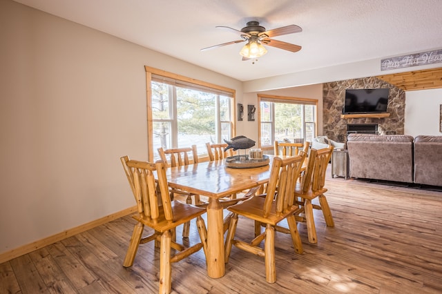 dining space with baseboards, a stone fireplace, light wood-style flooring, and a ceiling fan