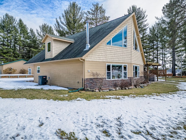 snow covered back of property featuring central air condition unit, stone siding, and a shingled roof