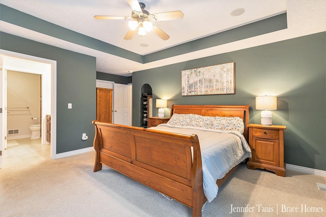 bedroom with a tray ceiling, light colored carpet, ensuite bath, and baseboards