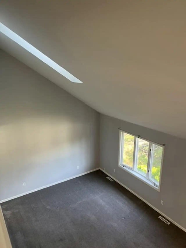 spare room with dark colored carpet, visible vents, vaulted ceiling with skylight, and baseboards