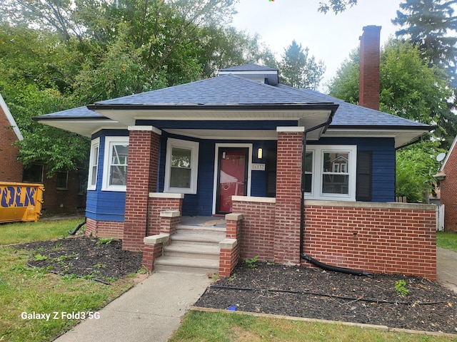 bungalow featuring a porch, brick siding, a shingled roof, and a chimney