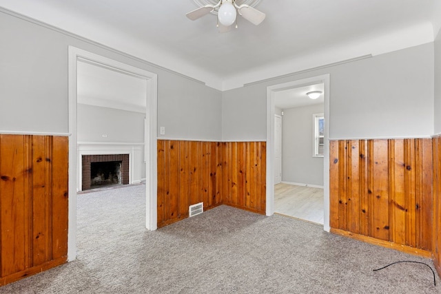 unfurnished living room with wooden walls, a wainscoted wall, carpet floors, ceiling fan, and a brick fireplace