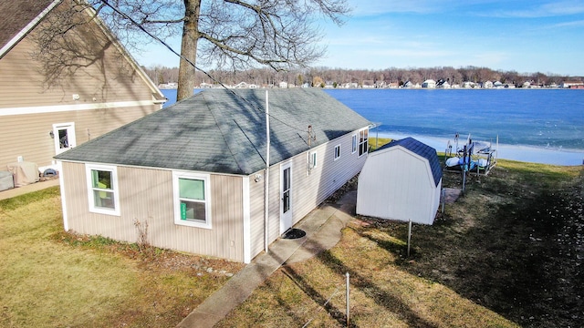 view of side of home with an outbuilding, roof with shingles, a storage unit, a water view, and a lawn