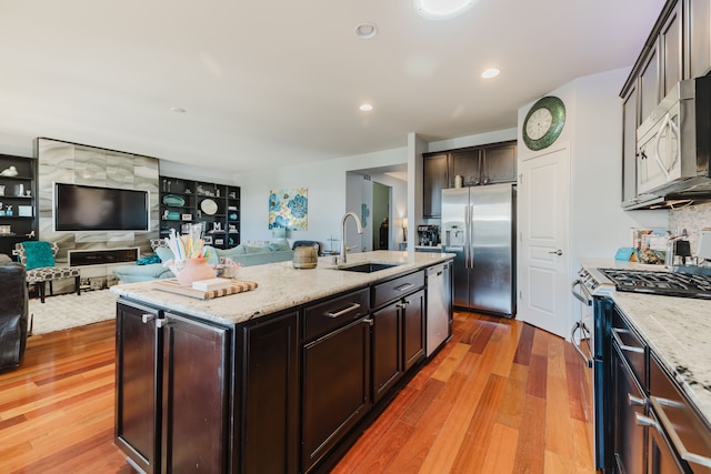 kitchen with light wood-type flooring, a kitchen island with sink, a sink, dark brown cabinetry, and appliances with stainless steel finishes