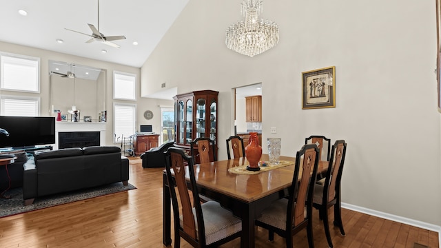 dining area featuring baseboards, hardwood / wood-style floors, ceiling fan with notable chandelier, a fireplace, and a high ceiling