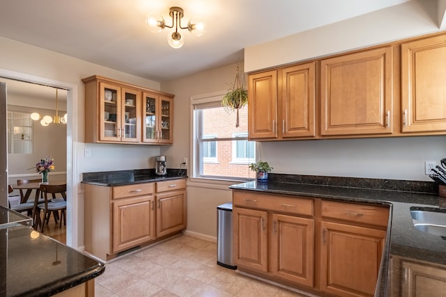 kitchen featuring glass insert cabinets, an inviting chandelier, brown cabinetry, and dark stone counters