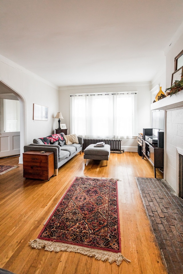 living room featuring radiator, light wood finished floors, arched walkways, a tile fireplace, and crown molding