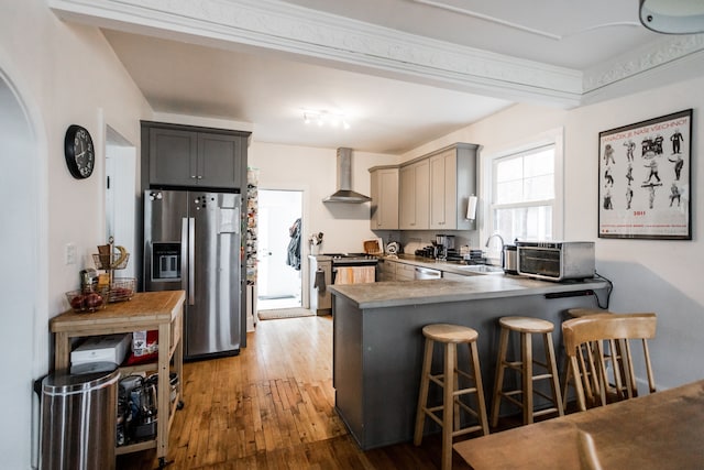 kitchen with a breakfast bar area, a peninsula, light wood-style flooring, stainless steel appliances, and wall chimney range hood