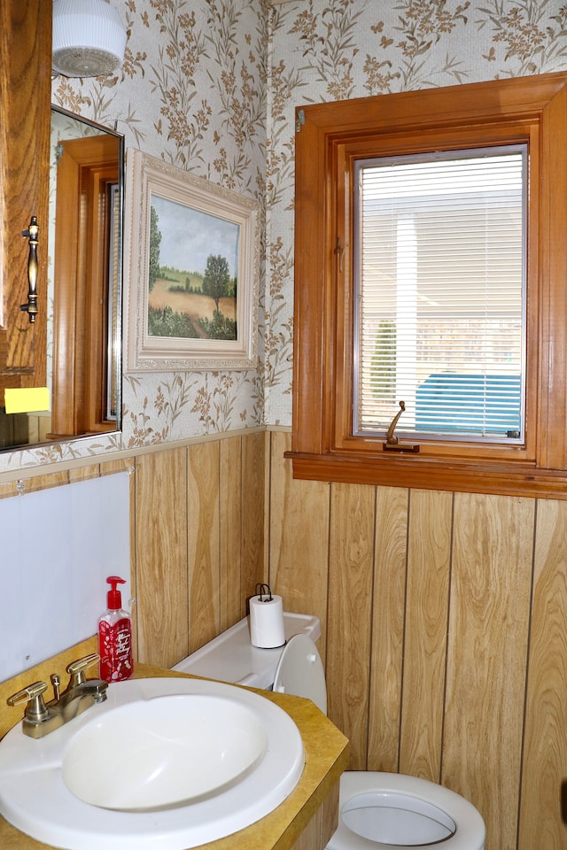 bathroom with wainscoting, toilet, wallpapered walls, and a sink