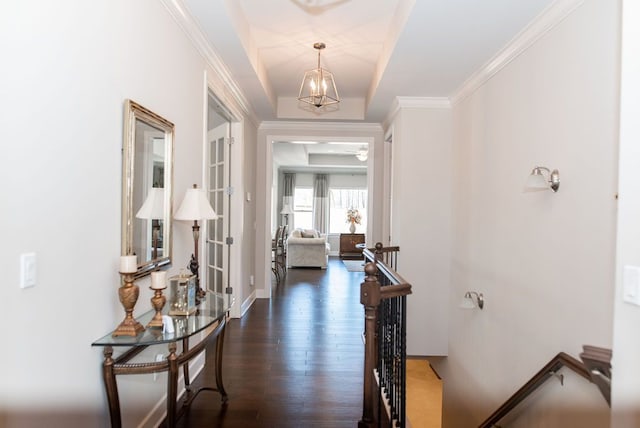 corridor with a tray ceiling, dark wood-type flooring, crown molding, a notable chandelier, and an upstairs landing