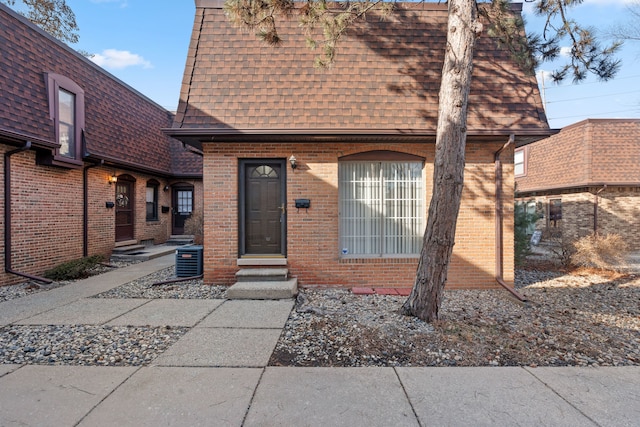 view of front facade featuring brick siding, a shingled roof, entry steps, central AC unit, and mansard roof