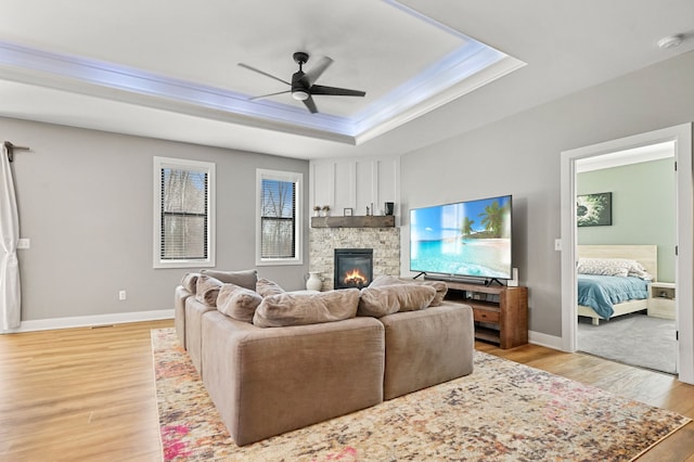 living room featuring a stone fireplace, a ceiling fan, a raised ceiling, and light wood-style floors