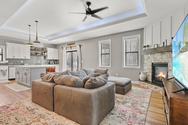 living room with light wood finished floors, baseboards, ceiling fan, a stone fireplace, and a raised ceiling