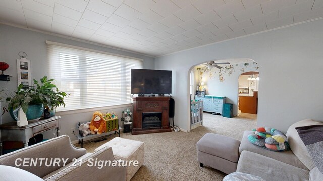 living area featuring carpet floors, a fireplace with raised hearth, arched walkways, ceiling fan, and ornamental molding