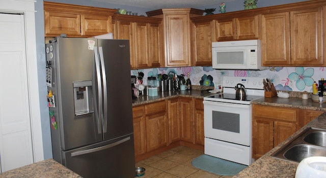 kitchen with brown cabinets, a sink, dark stone countertops, white appliances, and light tile patterned flooring