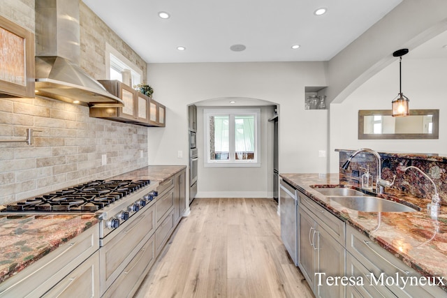 kitchen with light stone countertops, light wood-type flooring, stainless steel appliances, wall chimney exhaust hood, and a sink