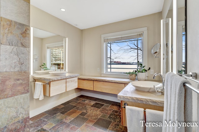 full bath featuring stone tile flooring, two vanities, a healthy amount of sunlight, and a sink