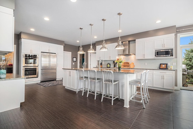 kitchen with wall chimney range hood, an island with sink, stainless steel appliances, white cabinetry, and backsplash