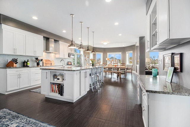 kitchen featuring wall chimney range hood, recessed lighting, white cabinets, and a center island with sink
