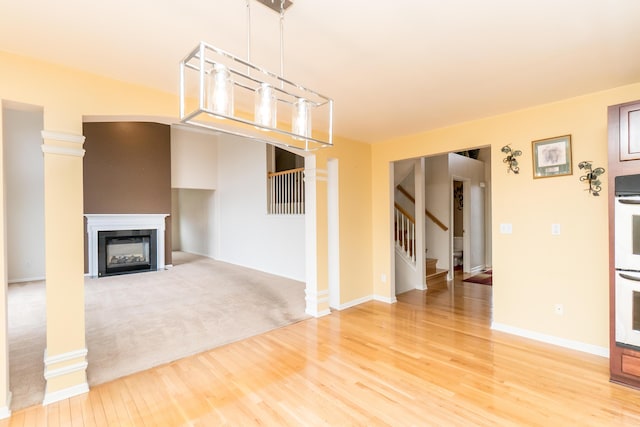 unfurnished living room featuring a glass covered fireplace, stairway, light wood-style floors, decorative columns, and baseboards