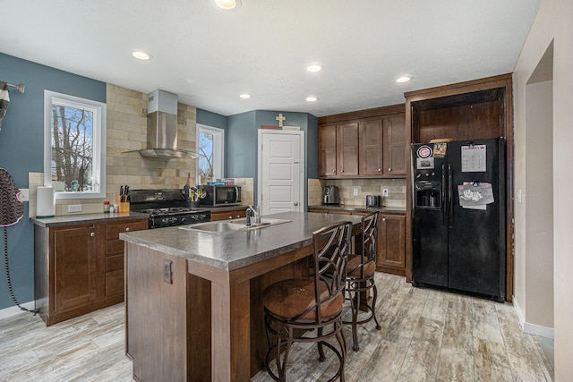 kitchen featuring dark countertops, light wood-style floors, black appliances, wall chimney exhaust hood, and a sink