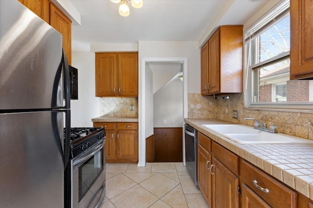 kitchen with tile countertops, light tile patterned floors, brown cabinets, appliances with stainless steel finishes, and a sink