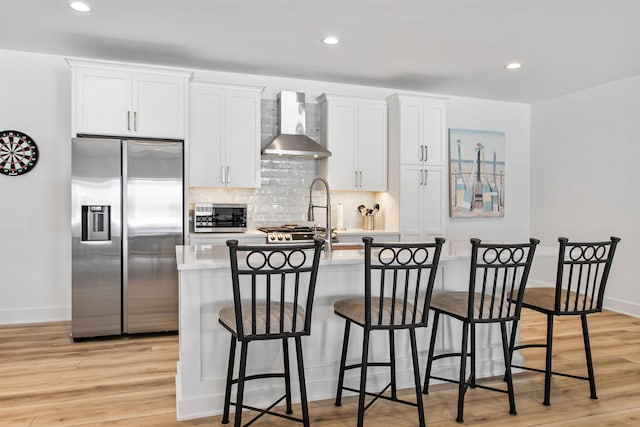 kitchen featuring a center island with sink, light wood-style flooring, appliances with stainless steel finishes, wall chimney range hood, and backsplash