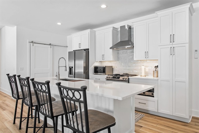 kitchen featuring wall chimney range hood, a breakfast bar area, an island with sink, a barn door, and appliances with stainless steel finishes