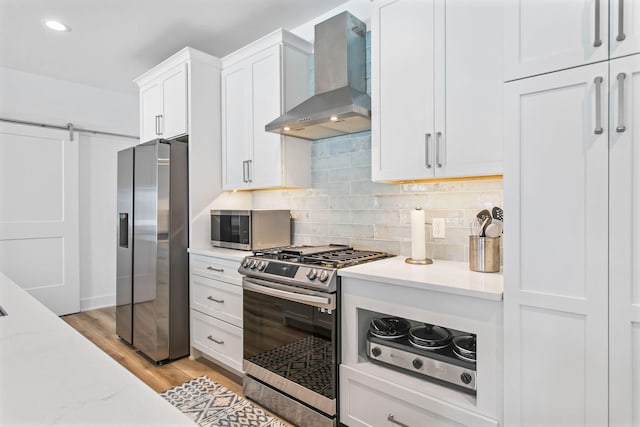 kitchen with white cabinetry, stainless steel appliances, a barn door, wall chimney range hood, and decorative backsplash