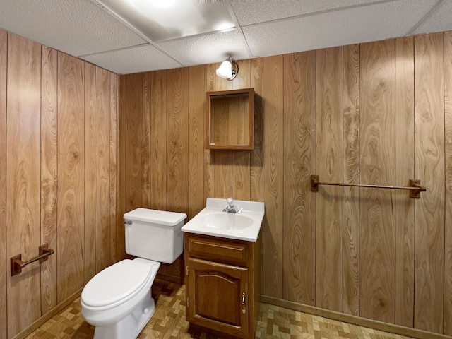 half bath with vanity, wooden walls, toilet, and a paneled ceiling