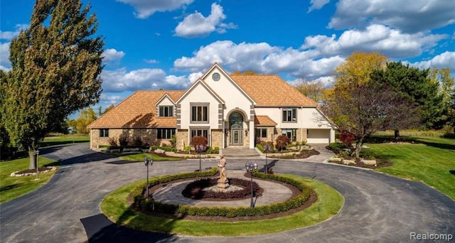 english style home with stucco siding, curved driveway, a front lawn, stone siding, and a tiled roof