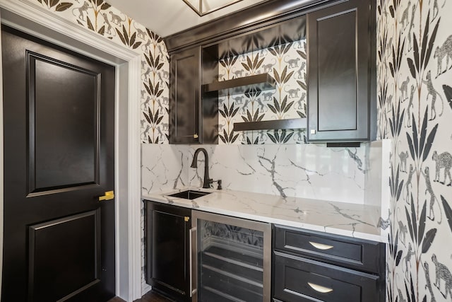 kitchen featuring light stone countertops, wallpapered walls, a sink, wine cooler, and tasteful backsplash