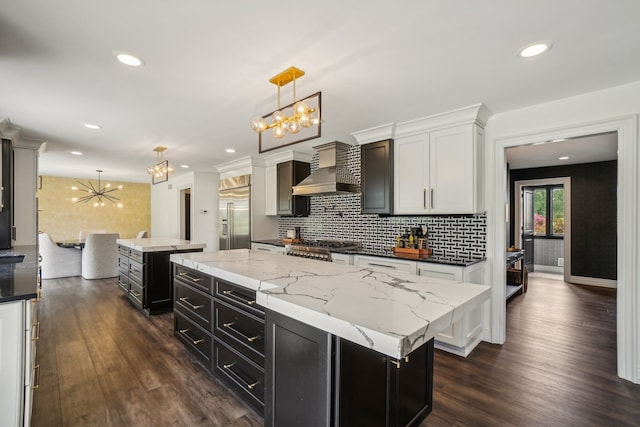 kitchen featuring an inviting chandelier, a center island, wall chimney range hood, dark cabinets, and stainless steel built in refrigerator