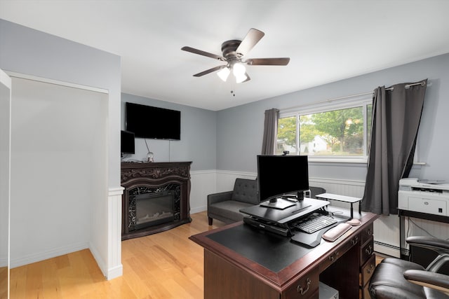 office space featuring ceiling fan, wainscoting, light wood-style flooring, a glass covered fireplace, and a baseboard radiator