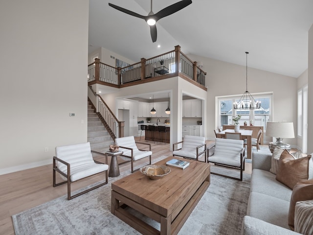 living area with baseboards, stairs, light wood-type flooring, ceiling fan with notable chandelier, and high vaulted ceiling