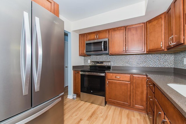 kitchen with brown cabinetry, light wood-style flooring, stainless steel appliances, decorative backsplash, and dark countertops