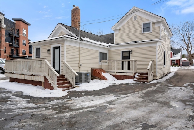 snow covered rear of property featuring central AC unit, a chimney, a deck, and roof with shingles