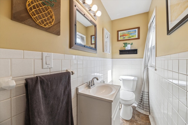 bathroom featuring toilet, stone finish flooring, tile walls, wainscoting, and vanity