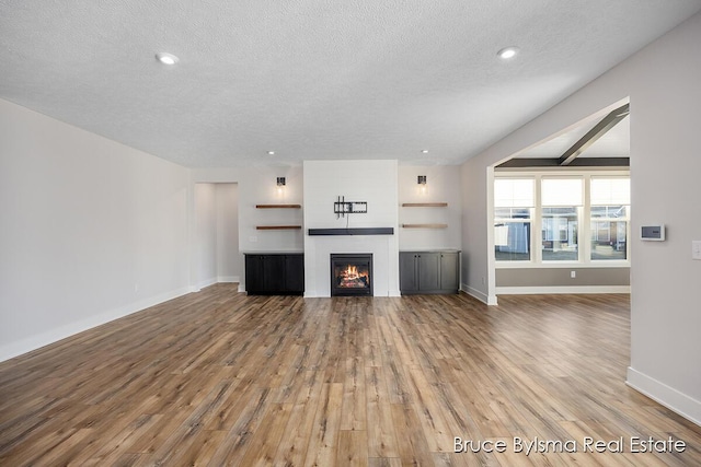 unfurnished living room featuring light wood-type flooring, a textured ceiling, a warm lit fireplace, recessed lighting, and baseboards