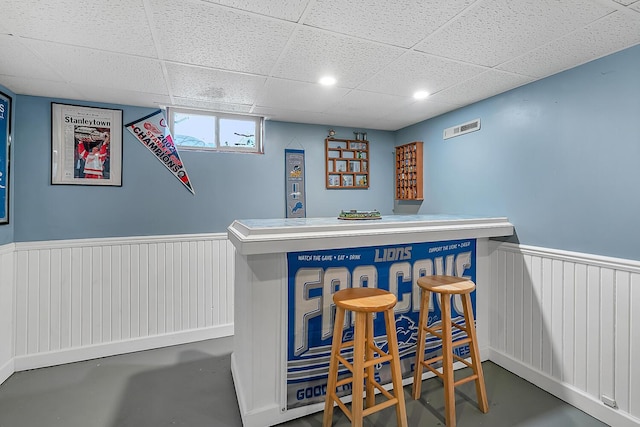 bar with a drop ceiling, a bar, concrete floors, and wainscoting