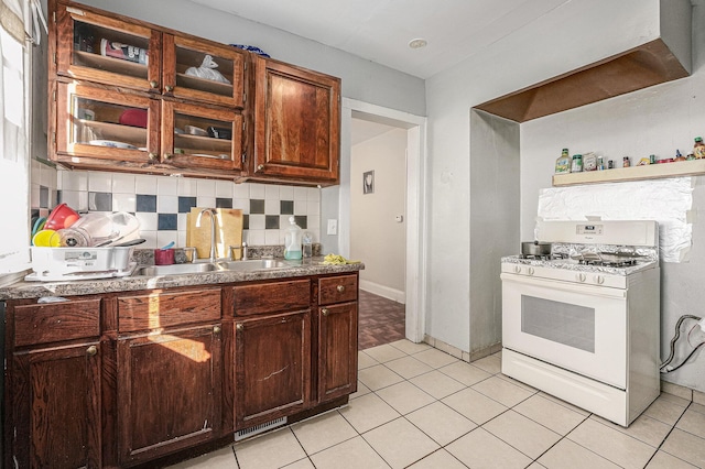 kitchen with white range with gas stovetop, light tile patterned flooring, a sink, glass insert cabinets, and tasteful backsplash