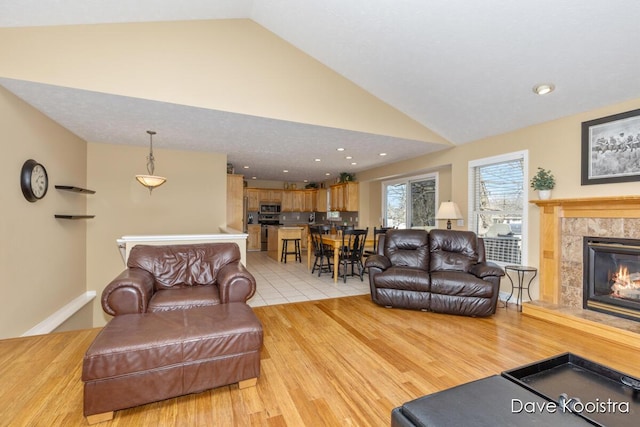 living area featuring baseboards, high vaulted ceiling, recessed lighting, a tiled fireplace, and light wood-type flooring