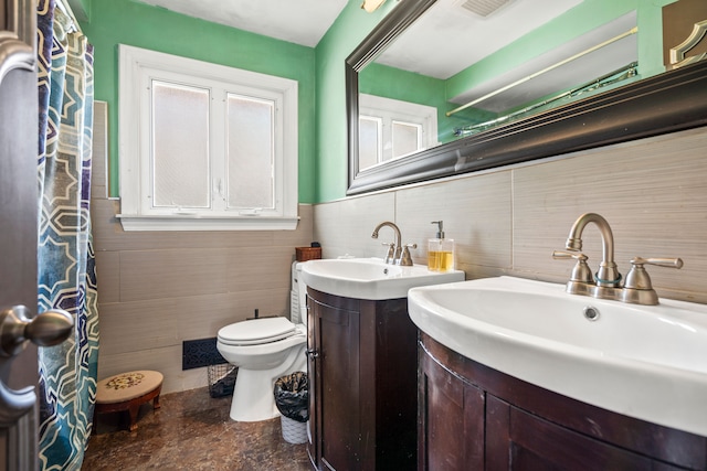 full bath featuring a wainscoted wall, two vanities, a sink, tile walls, and toilet