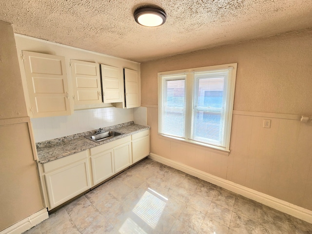 kitchen featuring a sink, a textured ceiling, white cabinetry, wainscoting, and light stone countertops