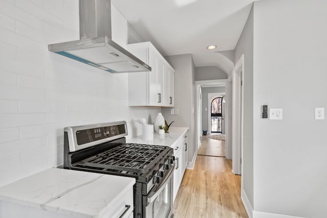 kitchen with wall chimney range hood, light stone counters, light wood-style flooring, stainless steel gas stove, and white cabinets