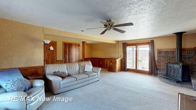 carpeted living area featuring a textured ceiling, a wood stove, a wainscoted wall, and ceiling fan