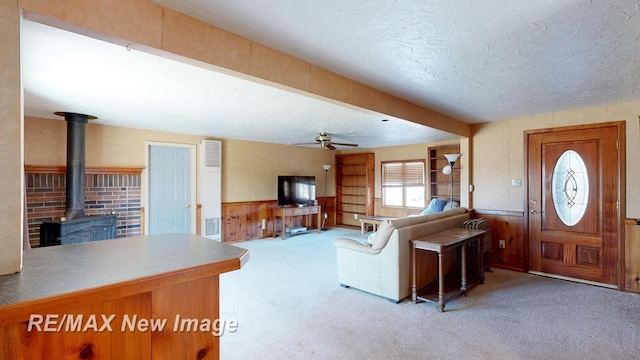 living room with a ceiling fan, carpet floors, a wood stove, wainscoting, and a textured ceiling